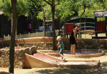 Un enfant et une femme jouent au mini-golf sous les arbres dans un hébergement glamping avec aire de jeux.