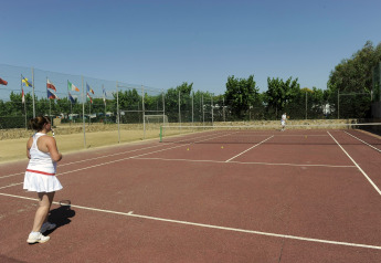 Twee personen spelen tennis op een buitenbaan bij een glamping, omringd door vlaggen en bomen.