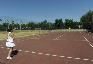 Deux personnes jouent au tennis sur un court extérieur dans un glamping, entourés de drapeaux et d’arbres.