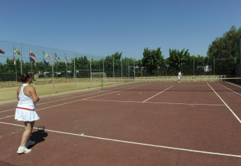 Dos personas juegan al tenis en una pista al aire libre en un glamping, rodeados de banderas y árboles.
