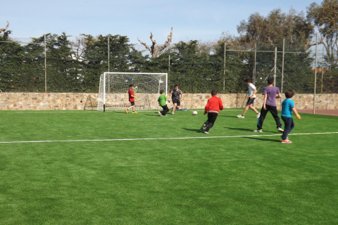 Niños y adultos juegan al fútbol en un campo verde en un alojamiento glamping, rodeados de naturaleza.