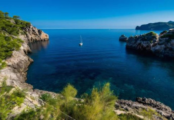 Stunning view of a peaceful cove with a sailboat, rocky cliffs, and blue waters near Camping del Mar BUNGALOW MEDITERRANI in Spain.