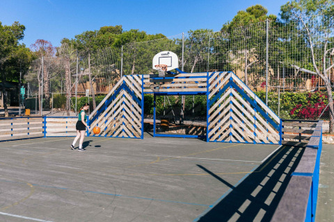 Cancha de baloncesto al aire libre en un glamping, cercada y rodeada de árboles, con una persona jugando.