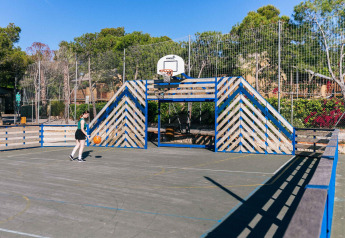 Cancha de baloncesto al aire libre en un glamping, cercada y rodeada de árboles, con una persona jugando.