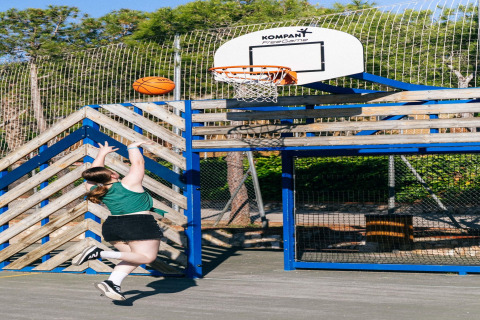 Una joven lanza una pelota de baloncesto a la canasta en una cancha al aire libre junto a un glamping.