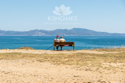 Two people sitting on a bench by the sea, enjoying mountain views at a scenic glamping accommodation.