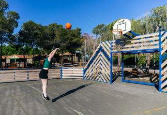 Vrouw gooit basketbal op een buitenplein met houten omheining bij een glamping accommodatie op zonnige dag.