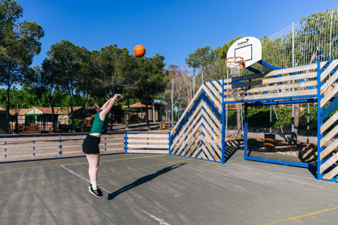Mujer lanzando a canasta en una pista exterior junto a una cerca de madera en un alojamiento glamping soleado.