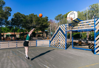 Mujer lanzando a canasta en una pista exterior junto a una cerca de madera en un alojamiento glamping soleado.