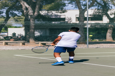 Un hombre juega al tenis en una pista al aire libre en un alojamiento glamping, con árboles y edificios de fondo.