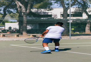Un hombre juega al tenis en una pista al aire libre en un alojamiento glamping, con árboles y edificios de fondo.