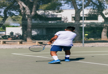 Un hombre juega al tenis en una pista al aire libre en un alojamiento glamping, con árboles y edificios de fondo.