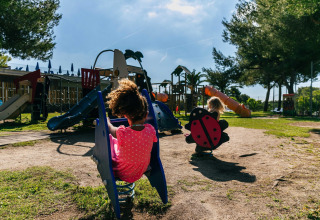 Niños juegan en un parque infantil de un alojamiento glamping, rodeados de árboles, toboganes y columpios.