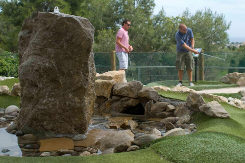 Dos hombres juegan minigolf junto a un arroyo artificial y rocas en un entorno natural de glamping.