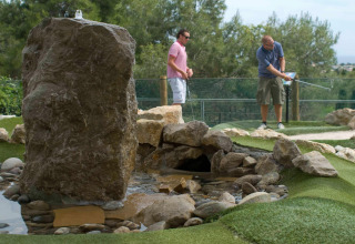 Dos hombres juegan minigolf junto a un arroyo artificial y rocas en un entorno natural de glamping.