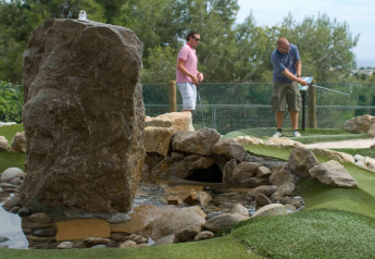 Dos hombres juegan minigolf junto a un arroyo artificial y rocas en un entorno natural de glamping.