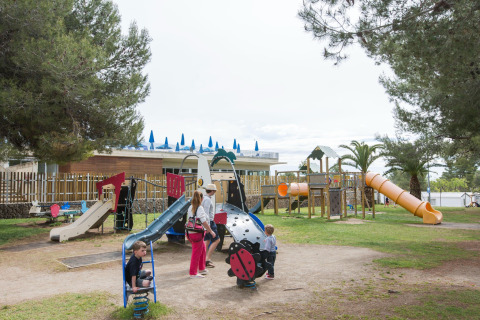 Niños y adultos jugando en un parque infantil en un resort de glamping, con árboles y toboganes.