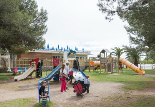 Niños y adultos jugando en un parque infantil en un resort de glamping, con árboles y toboganes.