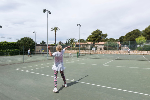 Mujer jugando al tenis en una pista exterior en un alojamiento glamping, rodeada de árboles y cabañas.