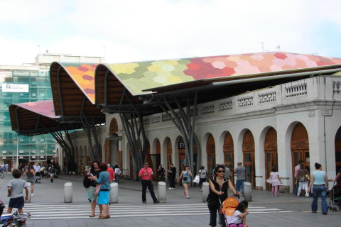 Personas alrededor de un mercado en España con techo ondulado colorido y una fachada blanca con arcos.