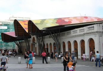 Personas alrededor de un mercado en España con techo ondulado colorido y una fachada blanca con arcos.