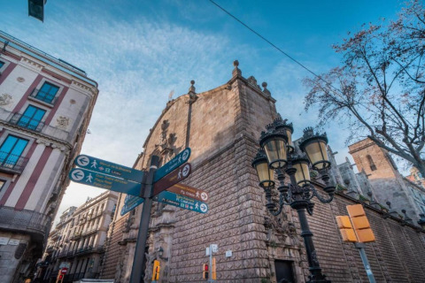 Señales peatonales y edificios antiguos en Barcelona, España, bajo cielo azul y farolas clásicas.