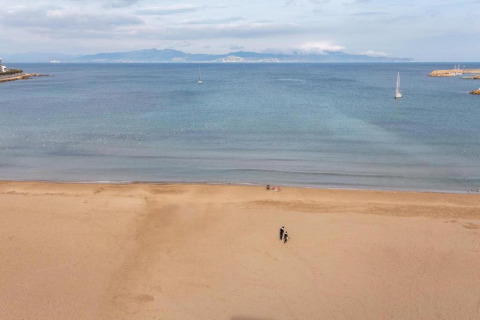 Beach scene at a glamping accommodation showing the sea, sailboats, and two people walking on sand.