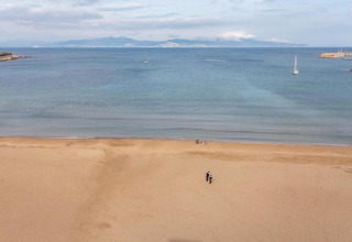 Beach scene at a glamping accommodation showing the sea, sailboats, and two people walking on sand.