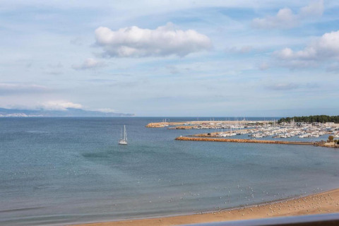 View from glamping accommodation of sandy beach, sailboat, and marina with yachts by the sea.