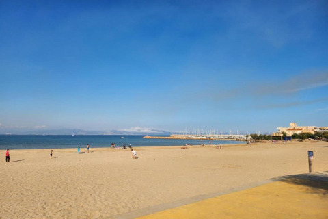 Photo of a sandy beach at a glamping spot, with few people, calm sea, blue sky, and marina distant.