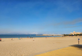 Photo of a sandy beach at a glamping spot, with few people, calm sea, blue sky, and marina distant.