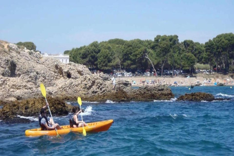 Two people kayaking in an orange kayak near rocky shores at Nomad Paraiso, Spain, with a beach in the background.