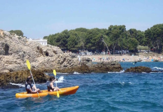 Two people kayaking in an orange kayak near rocky shores at Nomad Paraiso, Spain, with a beach in the background.