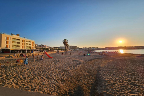 Sunset view at Nomad Paraiso beach in Spain, with people, a playground, loungers, and nearby buildings.