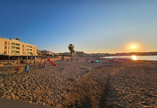 Sunset view at Nomad Paraiso beach in Spain, with people, a playground, loungers, and nearby buildings.