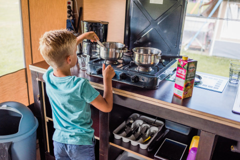 Jongen kookt op een gasvuur in een safaritentkeuken met potten, fruitsap en besteklade zichtbaar.