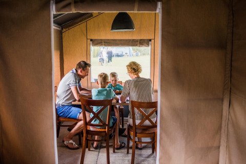 Een familie zit rond een tafel in een safaritent en geniet samen van een moment op de camping.