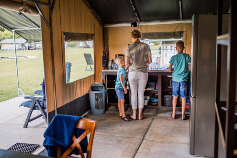 Binnen in een safaritent staan een vrouw met twee kinderen aan het aanrecht, omgeven door kampeergerief.