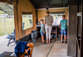 Binnen in een safaritent staan een vrouw met twee kinderen aan het aanrecht, omgeven door kampeergerief.