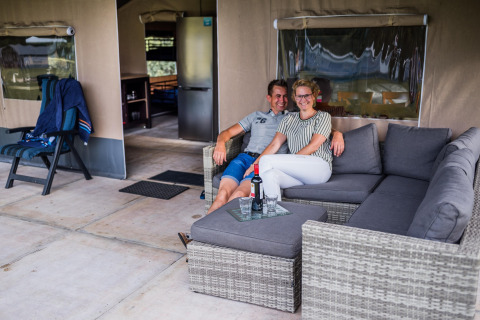 Couple détendu sur un canapé gris devant une tente safari avec bouteille de vin et verres sur la table.