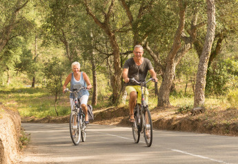 Zwei ältere Menschen fahren fröhlich mit dem Fahrrad auf einer Straße im Safari-Zelt Resort «Begur 1» in Spanien.