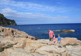 Eine Familie steht auf Felsen und blickt auf das Meer bei Resort «Begur 1» in Spanien unter blauem Himmel.
