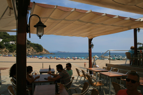 Blik vanuit een schaduwrijke tentcafé over het levendige strand met zeezicht in Begur, Spanje.