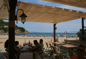Blick von einem schattigen Café mit Segeldach über den lebhaften Strand und das Meer in Begur, Spanien.