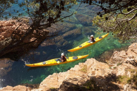 Two people kayak in bright yellow kayaks through clear turquoise waters, surrounded by rocky cliffs in Spain.