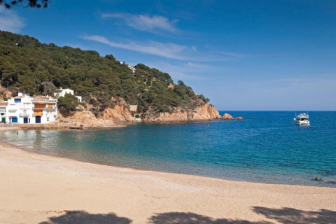 Belle plage au Resort Begur 3 en Espagne, avec falaises, pins, maisons blanches et un bateau sur la mer.