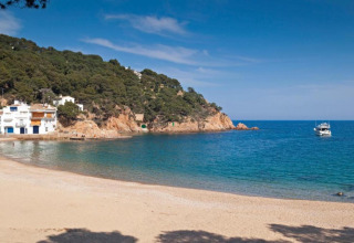 Belle plage au Resort Begur 3 en Espagne, avec falaises, pins, maisons blanches et un bateau sur la mer.