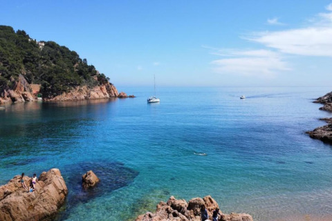 Belle vue côtière avec une mer bleue limpide, des bateaux, des rochers et des collines verdoyantes.