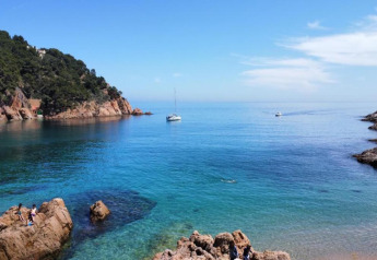 Belle vue côtière avec une mer bleue limpide, des bateaux, des rochers et des collines verdoyantes.