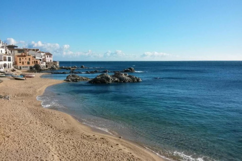 Plage de sable avec eau bleue limpide, rochers et maisons colorées sous un ciel bleu éclatant.